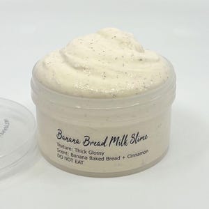 May include: A clear plastic jar filled with off-white, speckled slime. The label reads: Banana Bread Milk Slime, Texture: Thick Glossy, Scent: Banana Baked Bread + Cinnamon, DO NOT EAT. A separate clear plastic lid sits to the left of the jar.
