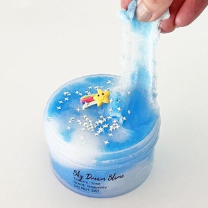 Op de afbeelding: Een blauwe en witte slime met een ster- en regenboogbedel in een doorzichtige container. De slime wordt uit de container getrokken en is erg rekbaar. De container heeft een label met de tekst "Sky Dream Slime" met de tekst "Textuur: Icee Geur: Heavenly NIET ETEN".