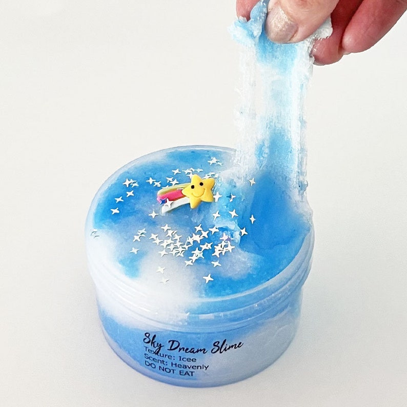 Op de afbeelding: Een blauwe en witte slime met een ster- en regenboogbedel in een doorzichtige container. De slime wordt uit de container getrokken en is erg rekbaar. De container heeft een label met de tekst "Sky Dream Slime" met de tekst "Textuur: Icee Geur: Heavenly NIET ETEN".