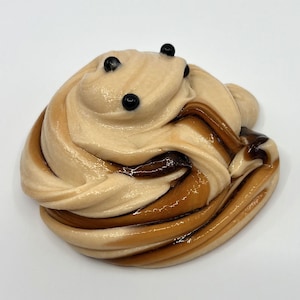 Puede incluir: Un remolino de slime beige y marr&oacute;n con tres cuentas negras en la parte superior. El slime brillante tiene una textura suave. Los colores y la disposici&oacute;n sugieren un tema de postre.