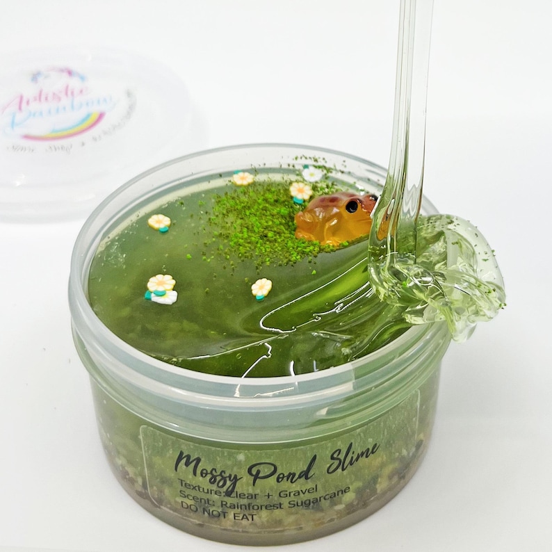 May include: A clear plastic container filled with green slime, decorated with small white flowers, green moss, and a small frog figurine. The label reads "Mossy Pond Slime". The slime is clear and has a clear and gravel texture.