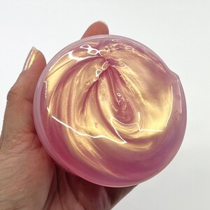 Peut inclure: Slime rose avec des tourbillons de paillettes dor&eacute;es. La slime est dans un r&eacute;cipient transparent et est tenue dans une main.