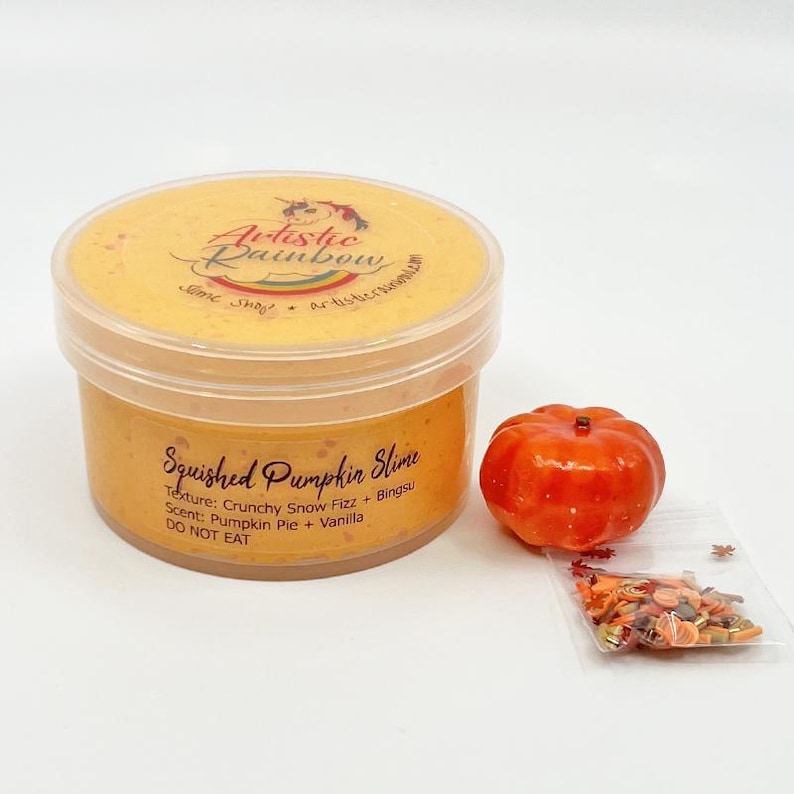 Op de afbeelding: Een pot oranje slijm met het opschrift "Squished Pumpkin Slime" en de tekst "Artistic Rainbow". Daarnaast een kleine oranje pompoen en een zakje herfst-thema sprinkles. De textuur is "Crunchy Snow Fizz".