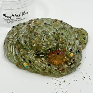 May include: A close-up of a green, clear slime with small gravel and a small, orange frog figurine. The slime has the text "Mossy Pond Slime" and "DO NOT EAT" on the container. The slime is a unique sensory toy.