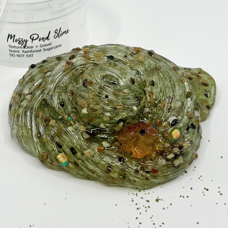 May include: A close-up of a green, clear slime with small gravel and a small, orange frog figurine. The slime has the text "Mossy Pond Slime" and "DO NOT EAT" on the container. The slime is a unique sensory toy.