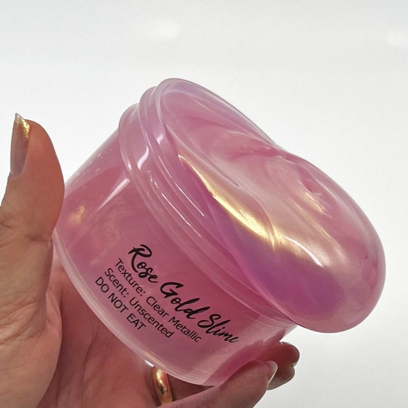 Peut inclure: Une slime rose transparente avec une brillance m&eacute;tallique dans un r&eacute;cipient en plastique. Le r&eacute;cipient porte une &eacute;tiquette sur laquelle est inscrit "Rose Gold Slime" avec le texte "Texture : M&eacute;tallique transparente, Parfum : Non parfum&eacute;, NE PAS MANGER".