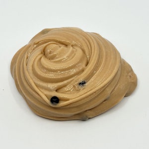 Puede incluir: Un remolino de slime marr&oacute;n claro con un acabado brillante. El slime tiene una textura suave y est&aacute; salpicado de peque&ntilde;as cuentas de color azul oscuro o negro. El fondo es blanco liso.