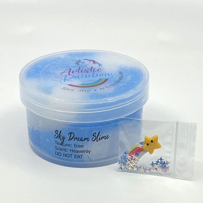 Op de afbeelding: Een doorzichtige plastic container gevuld met blauwe en witte slijm met het label "Sky Dream Slime". De slijm heeft een "Icee" textuur en een "Heavenly" geur. De container is gelabeld "Artistic Rainbow Slime Shop". Een kleine doorzichtige plastic zak met een gele ster charme en regenboog glitter is inbegrepen.