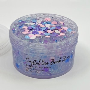 May include: A clear plastic jar filled with purple and blue tinted slime containing small shell-shaped charms in pink, purple, and white. The label reads: Crystal Sea Bomb Slime, Texture: Crunch Bomb, Scent: Unscented, DO NOT EAT.