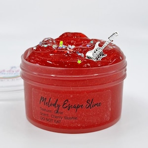 Puede incluir: Slime rojo con purpurina y un dije de guitarra plateado en un frasco transparente. La etiqueta dice "Melody Escape Slime", "Textura: Clara", "Aroma: Slush de cereza", y "No comer".