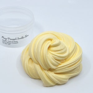 May include: A close-up of a yellow slime with a thick and glossy texture. The slime is in a clear container with a label that reads "Mango Pineapple Smoothie Slime". The label also includes the text "Texture: Thick & Glossy", "Scent: Mango Pineapple", and "DO NOT EAT".