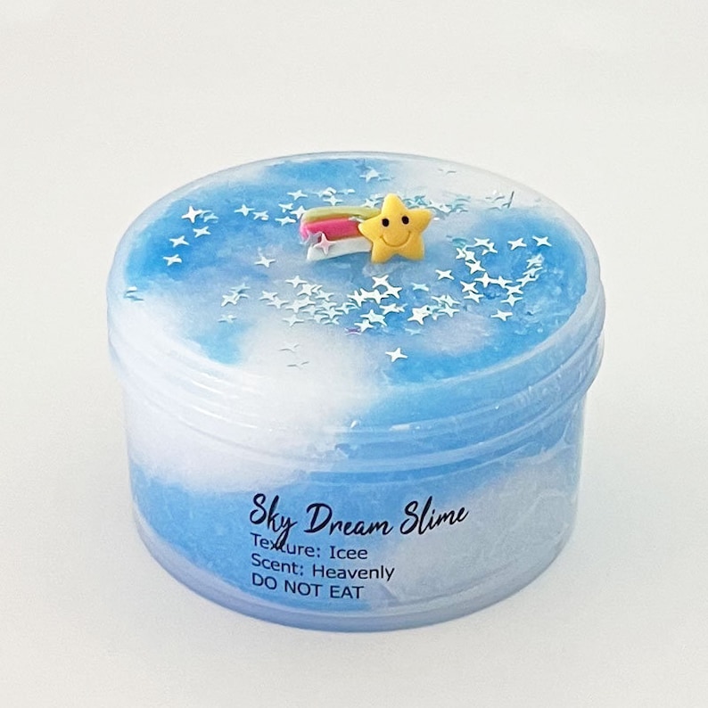 Op de afbeelding: Een blauwe en witte slime met een ster- en regenboogbedel in een doorzichtige plastic container. De slime is gelabeld met "Sky Dream Slime" met de tekst "Textuur: Icee, Geur: Hemels, NIET ETEN".