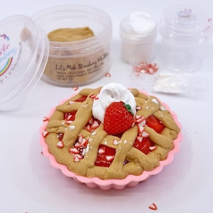 Puede incluir: Un kit de manualidades con temática de tarta de fresas, con un molde de silicona rosa. La tarta tiene una corteza de rejilla, relleno rojo, crema batida y una fresa. El kit incluye recipientes de crema de nube, arcilla de gelatina y masa de galletas.
