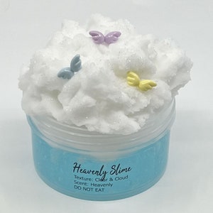 May include: White fluffy slime with glitter in a clear container with a blue lid. The slime has three small colorful wings on top. The container has the text "Heavenly Slime" with the words "Texture: Clear & Cloud" and "Scent: Heavenly" below. The text "DO NOT EAT" is at the bottom of the container.