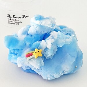 Op de afbeelding: Blauwe en witte slijm met een gele ster en een regenboogbedel. De slijm is gelabeld "Sky Dream Slime" met de tekst "Texture: Icee Scent: Heavenly DO NOT EAT".