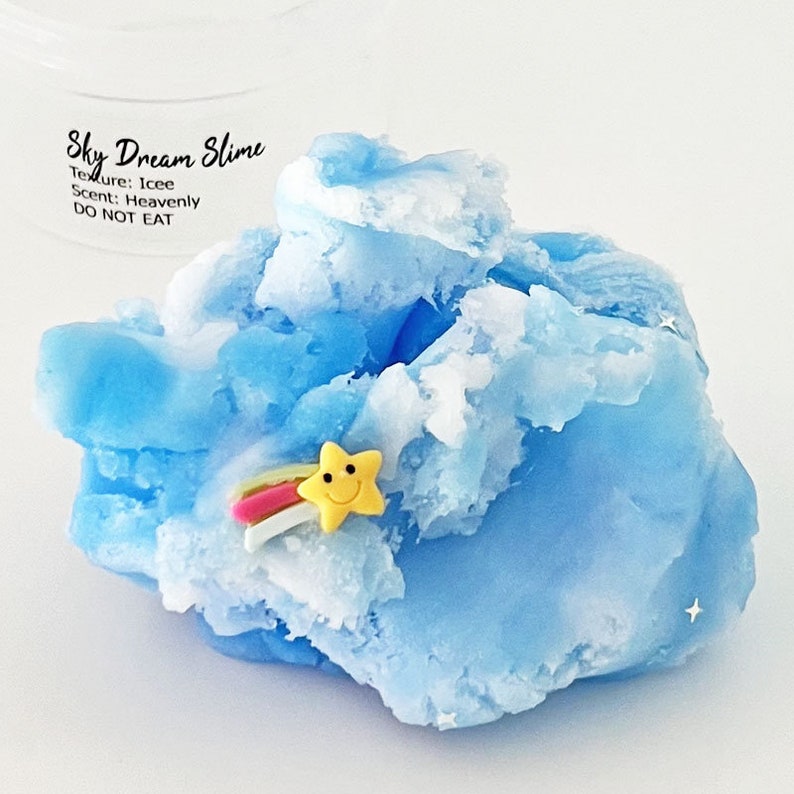 Op de afbeelding: Blauwe en witte slijm met een gele ster en een regenboogbedel. De slijm is gelabeld "Sky Dream Slime" met de tekst "Texture: Icee Scent: Heavenly DO NOT EAT".