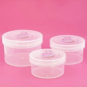 May include: Three clear plastic jars with white lids are arranged on a pink background. Each jar has a circular label on the lid with the text "Artistic Rainbow". The jars are different sizes, with the largest on the left.