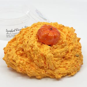 Op de afbeelding: Oranjegekleurde slijm met een kleine, oranje pompoen erop. De slijm heeft een textuur, knapperige uitstraling. De container naast de slijm leest "Squished Pumpkin Slime" met de textuur en geur.