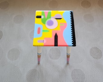 May include: A small, colorful stool with a geometric design painted on the top. The design features a mix of yellow, pink, blue, green, and black colors.