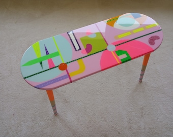 Könnte beinhalten: Ein rosa ovaler Couchtisch mit bunten geometrischen Mustern und orange-weißen gestreiften Beinen.