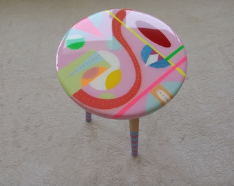 Pode incluir: Um banquinho redondo rosa com três pernas. A parte superior do banquinho é pintada com um design abstrato colorido que apresenta formas geométricas, linhas e pontos em tons de rosa, vermelho, amarelo, verde, azul e branco.
