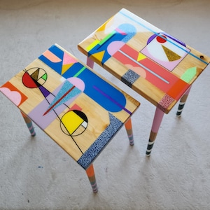 May include: Two small wooden tables with vibrant geometric designs. The tabletops display a mix of shapes and colours, including pink, blue, yellow, and black. The legs are painted with stripes and solid colours. Each table is approximately 61 cm long.