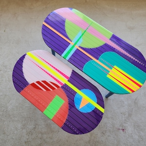 May include: Two oval-shaped tables with purple, pink, green, yellow, and red geometric patterns painted on the surface. The tables are made of wood and have a distressed look.