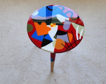 May include: A small, round table with a wooden leg. The tabletop is painted with a colorful abstract design featuring red, blue, orange, yellow, black, white, and gray.