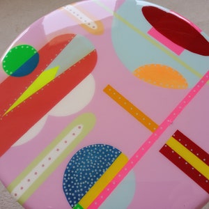 May include: A pink round table top with a colorful geometric design. The design features circles, lines, and dots in various colors, including red, yellow, blue, green, and white.