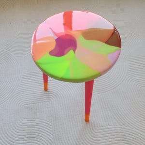 Puede incluir: Una pequeña mesa redonda con un diseño abstracto y colorido. La tapa presenta un remolino de tonos rosa, naranja, verde y morado. Las tres patas son de color rosa brillante con puntas naranjas. La mesa es una pieza decorativa.