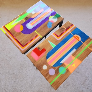 Könnte beinhalten: Zwei Holz-Beistelltische mit geometrischen Mustern, die in leuchtenden Farben wie Rosa, Blau, Grün, Gelb und Lila bemalt sind. Die Tische haben schwarze Metallbeine.