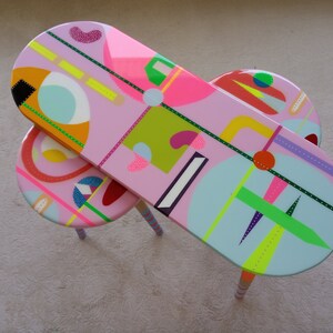 May include: Two oval-shaped nesting tables with pink, blue, green, yellow, and orange geometric patterns. The tables have wooden legs with pink, blue, and white stripes.