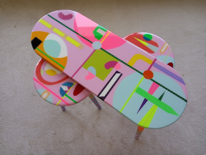 May include: Two oval-shaped nesting tables with pink, blue, green, yellow, and orange geometric patterns. The tables have wooden legs with pink, blue, and white stripes.
