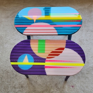 May include: Two oval-shaped wooden tables with colourful geometric designs. The top table is blue with pink, yellow, and green accents. The bottom table is purple with pink, yellow, green, and red accents.