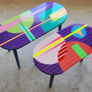 May include: Two oval-shaped side tables with purple, pink, green, blue and yellow geometric patterns painted on the top. The tables have blue legs.