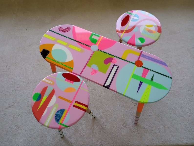 May include: A set of three pink oval-shaped tables with colorful geometric designs and black, white, and green accents. The tables have wooden legs.