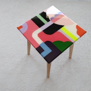 Op de afbeelding: Een kleine vierkante tafel met houten poten en een kleurrijk abstract ontwerp op de bovenkant. Het ontwerp bevat geometrische vormen in roze, groen, blauw, geel en zwart.