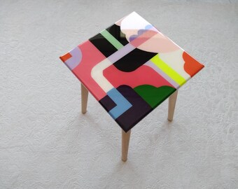 May include: A small square table with wooden legs and a colorful abstract design on the top. The design features geometric shapes in pink, green, blue, yellow, and black.