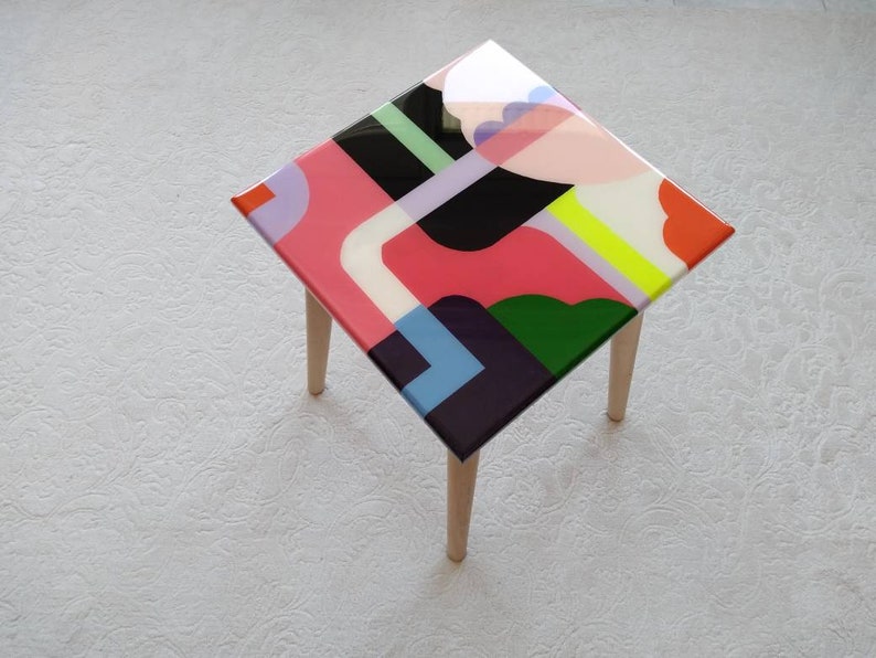 Op de afbeelding: Een kleine vierkante tafel met houten poten en een kleurrijk abstract ontwerp op de bovenkant. Het ontwerp bevat geometrische vormen in roze, groen, blauw, geel en zwart.