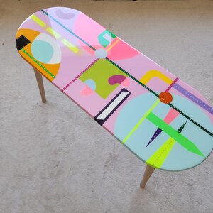 K&ouml;nnte beinhalten: Ein ovaler Couchtisch in Pink mit geometrischen Formen und bunten Mustern, die auf die Tischplatte gemalt sind. Der Tisch hat Holzbeine.