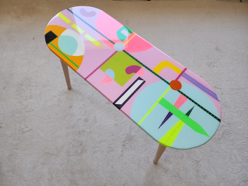 K&ouml;nnte beinhalten: Ein ovaler Couchtisch in Pink mit geometrischen Formen und bunten Mustern, die auf die Tischplatte gemalt sind. Der Tisch hat Holzbeine.
