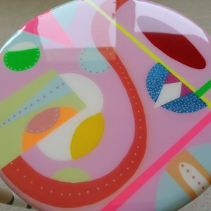 K&ouml;nnte beinhalten: Ein runder rosa Hocker mit einem farbenfrohen abstrakten Design. Das Design zeigt geometrische Formen, Punkte und Linien in verschiedenen Farben, darunter Rot, Gelb, Gr&uuml;n, Blau und Wei&szlig;.
