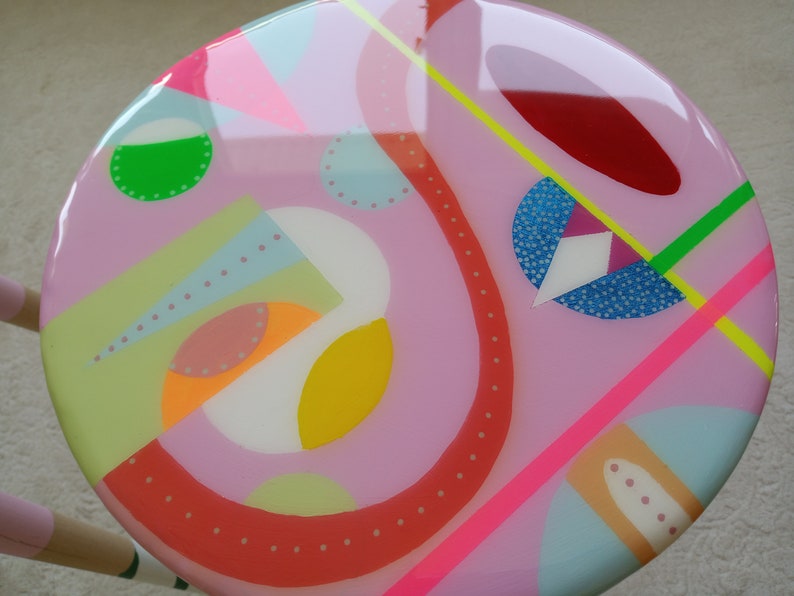 K&ouml;nnte beinhalten: Ein runder rosa Hocker mit einem farbenfrohen abstrakten Design. Das Design zeigt geometrische Formen, Punkte und Linien in verschiedenen Farben, darunter Rot, Gelb, Gr&uuml;n, Blau und Wei&szlig;.