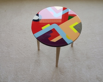 May include: A small, round side table with a colorful, abstract design on the tabletop. The table features a glossy finish with a red, blue, yellow, and purple geometric pattern. It stands on three pale wooden legs.