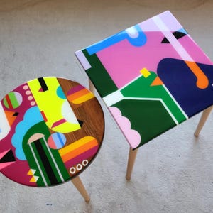 May include: Two colourful geometric patterned side tables. One table is round with a pink, blue, yellow, green, and black design. The other table is square with a pink, green, blue, and orange design.