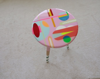 May include: A pink round table with three legs, painted with a colorful geometric design. The table top features circles, lines, and abstract shapes in shades of red, blue, yellow, green, and white.