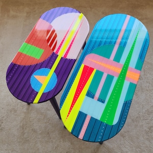 May include: Two oval-shaped tables with colourful geometric patterns. The table on the left has a purple, red, green, and yellow design. The table on the right has a blue, green, pink, and yellow design.