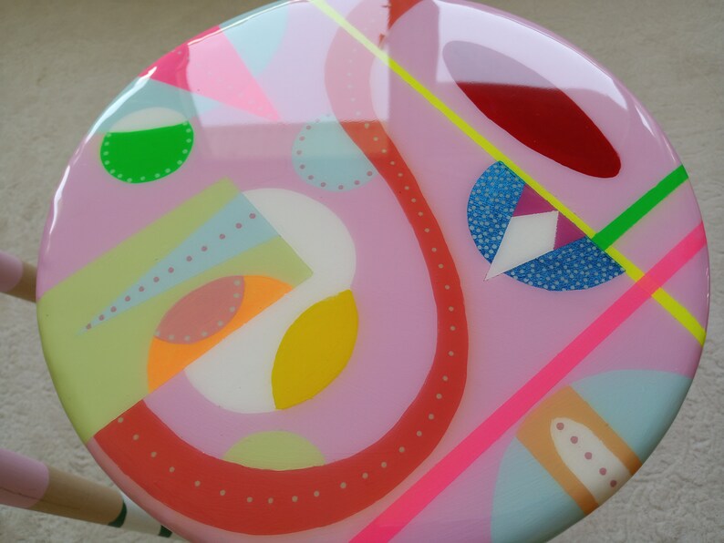 May include: A pink round stool with a colorful abstract design. The design features geometric shapes, dots, and lines in various colors, including red, yellow, green, blue, and white.