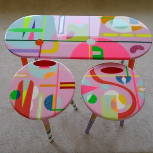 May include: A set of three pink painted wooden furniture pieces with geometric patterns in bright colors. The set includes a table and two stools.