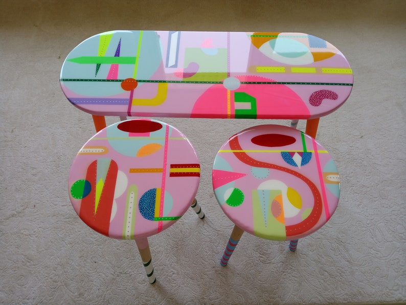 May include: A set of three pink painted wooden furniture pieces with geometric patterns in bright colors. The set includes a table and two stools.